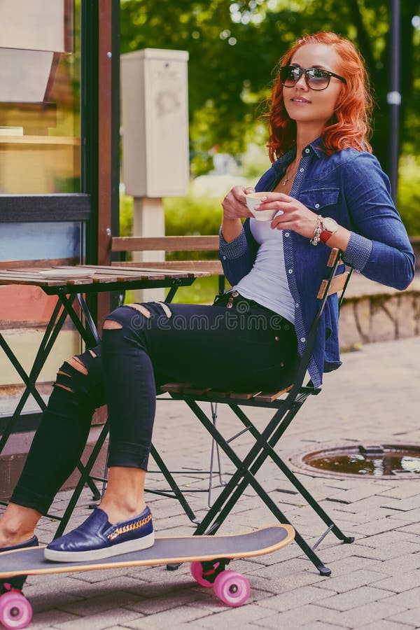 La ragazza della testarossa beve il caffè dopo avere pattinato su Longboard immagini stock