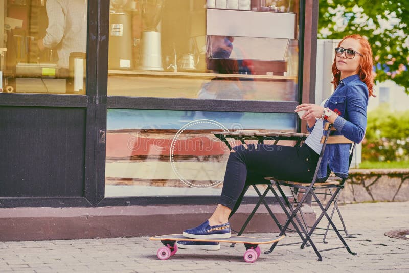 La ragazza della testarossa beve il caffè dopo avere pattinato su Longboard fotografia stock libera da diritti
