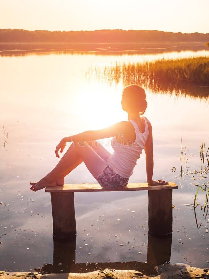 La ragazza gode di bello tramonto che si siede su un banco dal fiume Ragazza che si siede vicino all'acqua all'aperto Lago dorato fotografia stock libera da diritti La ragazza gode di bello tramonto che si siede su un banco dal fiume Ragazza che si siede vicino all'acqua all'aperto Lago dorato fotografia stock libera da diritti