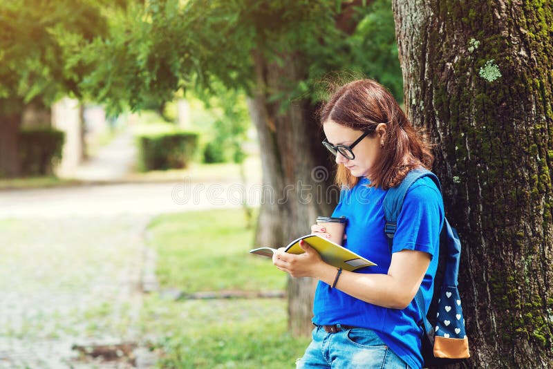 La ragazza graziosa dello studente in vetri, legge il taccuino, beve il caffè nel parco della città universitaria Vacanze estive, fotografie stock La ragazza graziosa dello studente in vetri, legge il taccuino, beve il caffè nel parco della città universitaria Vacanze estive, fotografie stock