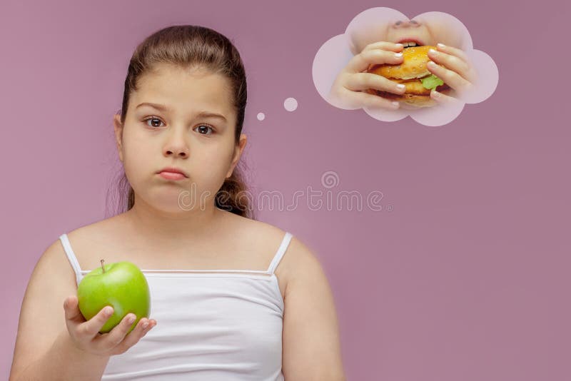 La ragazza mangia Apple verde, ma i sogni circa l'hamburger Alimento armonioso e sano per i bambini Bambino che mangia spuntino s immagini stock libere da diritti La ragazza mangia Apple verde, ma i sogni circa l'hamburger Alimento armonioso e sano per i bambini Bambino che mangia spuntino s immagini stock libere da diritti