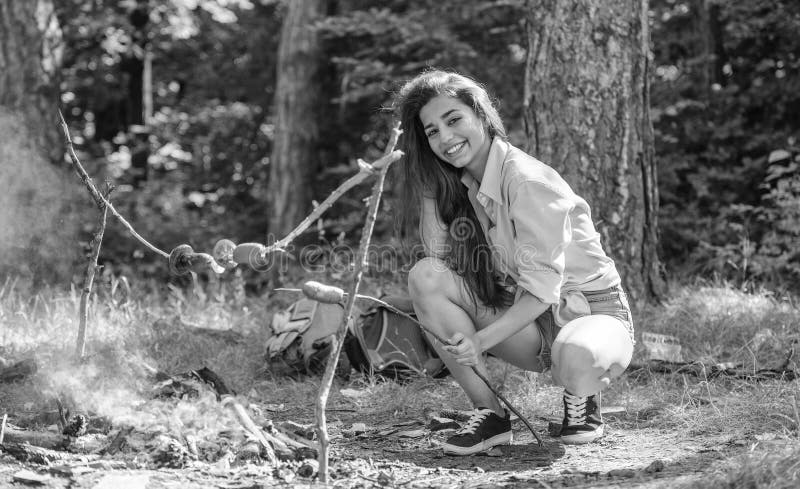 La ragazza si siede vicino alla salsiccia della torrefazione del falò sul bastone Turista della donna che produce alimento sopra fotografia stock La ragazza si siede vicino alla salsiccia della torrefazione del falò sul bastone Turista della donna che produce alimento sopra fotografia stock