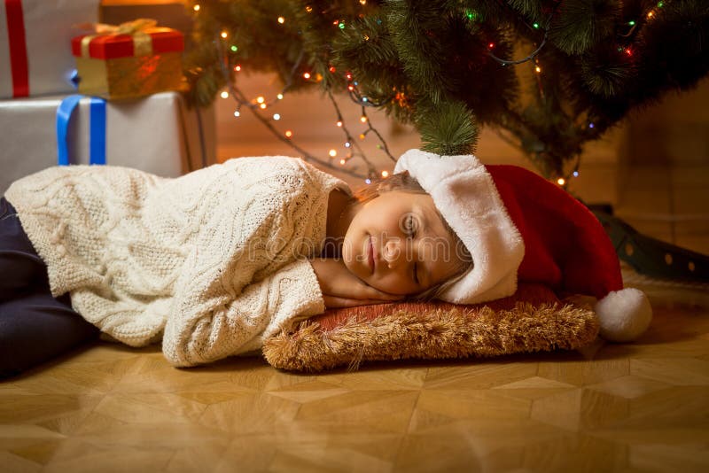 La ragazza sveglia in cappello di Santa è caduto addormentato sotto l'albero di Natale fotografia stock libera da diritti La ragazza sveglia in cappello di Santa è caduto addormentato sotto l'albero di Natale fotografia stock libera da diritti