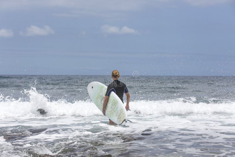 La resaca entra en el agua Persona que practica surf de sexo masculino que entra en el mar con su tablero en un traje que practic imágenes de archivo libres de regalías La resaca entra en el agua Persona que practica surf de sexo masculino que entra en el mar con su tablero en un traje que practic imágenes de archivo libres de regalías