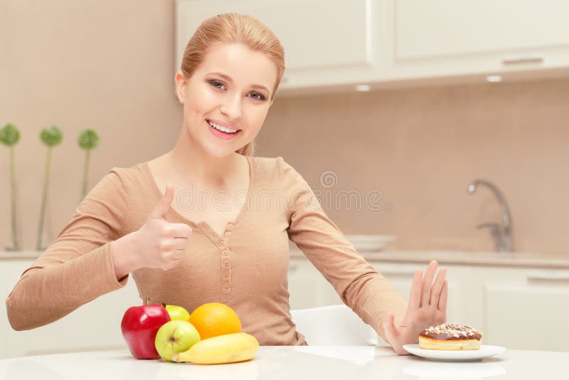 Made the right choice. Young lady sits at the table showing thumb up because she has strong will to decline eating donut and prefers apples and oranges. Made the right choice. Young lady sits at the table showing thumb up because she has strong will to decline eating donut and prefers apples and oranges