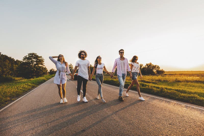 La société de jeunes types élégants heureux marchent sur une route de campagne un jour ensoleillé image stock La société de jeunes types élégants heureux marchent sur une route de campagne un jour ensoleillé image stock