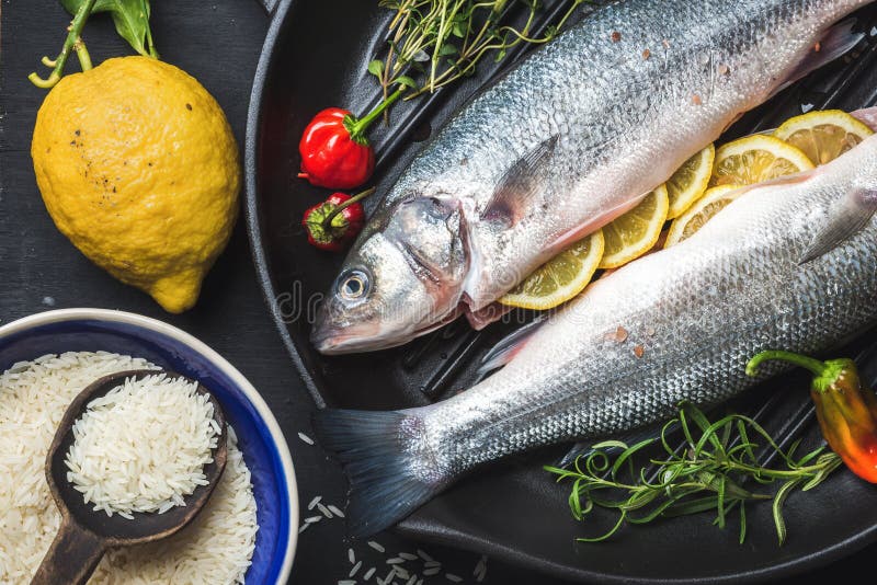 La spigola cruda cruda con riso, il limone, le erbe e le spezie sul grigliare nero rivestono di ferro la pentola sopra fondo scur fotografia stock libera da diritti La spigola cruda cruda con riso, il limone, le erbe e le spezie sul grigliare nero rivestono di ferro la pentola sopra fondo scur fotografia stock libera da diritti