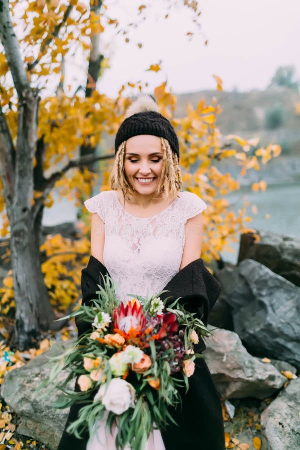 La sposa alla moda con un mazzo rustico sta posando prima di un lago sulla collina Giovane bionda in un cappello tricottato con i immagini stock libere da diritti La sposa alla moda con un mazzo rustico sta posando prima di un lago sulla collina Giovane bionda in un cappello tricottato con i immagini stock libere da diritti