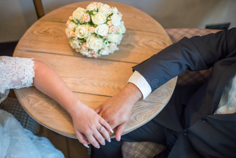 La sposa e lo sposo tengono le mani di ciascuno tabella di seduta Vicino al mazzo di nozze immagine stock libera da diritti La sposa e lo sposo tengono le mani di ciascuno tabella di seduta Vicino al mazzo di nozze immagine stock libera da diritti