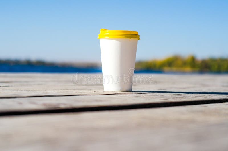 La taza del Libro Blanco con una tapa plástica amarilla se coloca en los tableros en la orilla del lago imagen de archivo libre de regalías La taza del Libro Blanco con una tapa plástica amarilla se coloca en los tableros en la orilla del lago imagen de archivo libre de regalías