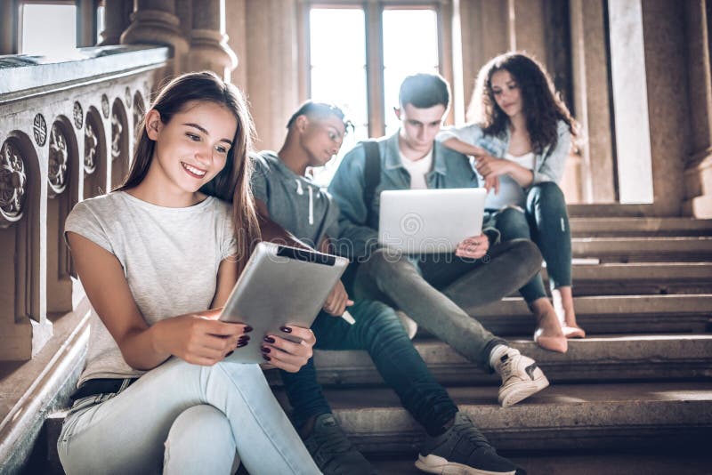 College life.A beautiful young student sitting on stairs in campus using the tablet while her friends work with laptop. College life.A beautiful young student sitting on stairs in campus using the tablet while her friends work with laptop.