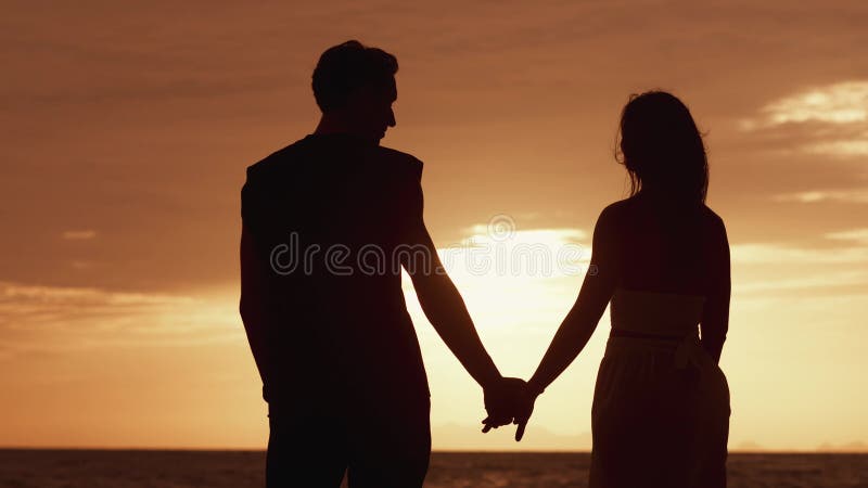 La visión trasera del hombre enamorado da la mano a una mujer en una cita contra una puesta de sol naranja. silueta de una pareja