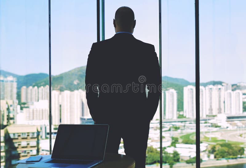La vue arrière d'un entrepreneur sûr d'homme regarde dans la grande fenêtre de bureau image stock La vue arrière d'un entrepreneur sûr d'homme regarde dans la grande fenêtre de bureau image stock