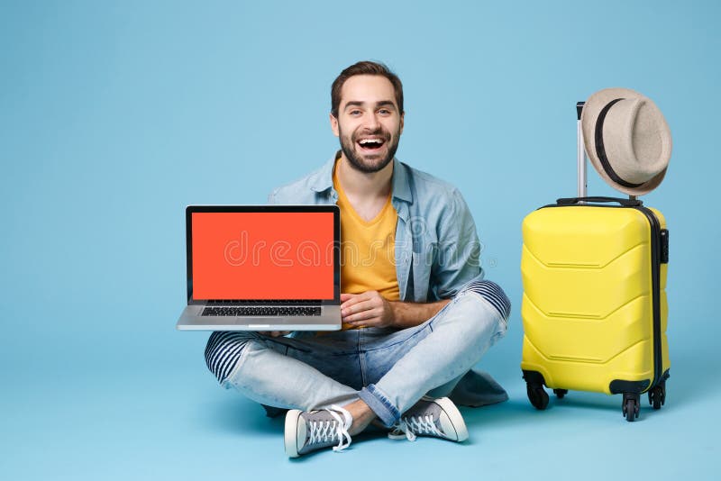 Laughing traveler tourist man in yellow clothes isolated on blue background. Passenger traveling abroad on weekend. Air flight journey concept. Sit near suitcase, hold laptop with blank empty screen. Laughing traveler tourist man in yellow clothes isolated on blue background. Passenger traveling abroad on weekend. Air flight journey concept. Sit near suitcase, hold laptop with blank empty screen