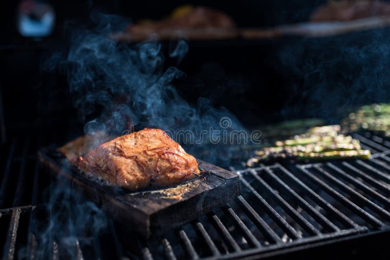 Image of a grill with salmon filet on wooden plank. Smoke is coming out of the grill boosting the fish smoky flavour. Image of a grill with salmon filet on wooden plank. Smoke is coming out of the grill boosting the fish smoky flavour