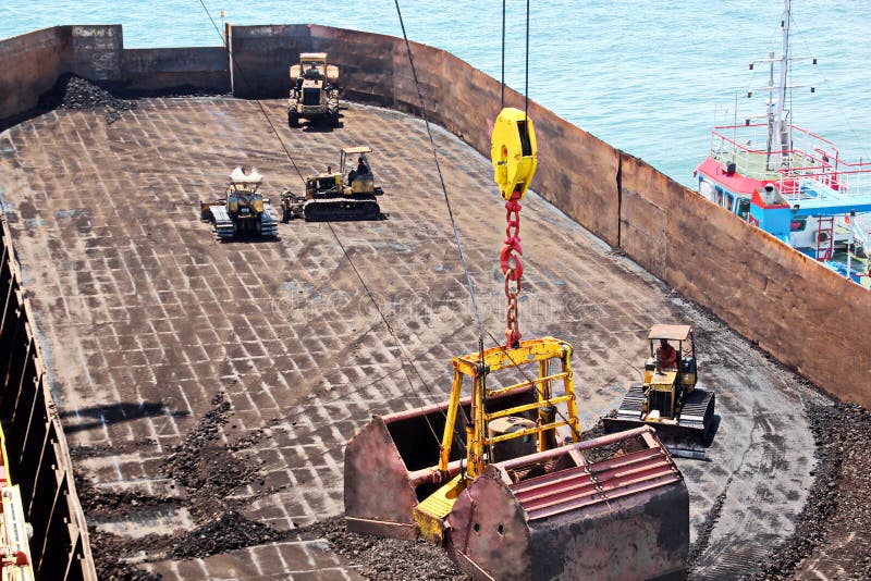 Loading coal from cargo barges onto a bulk carrier using ship cranes and grabs at the port of Samarinda, Indonesia. View of a close-up of the work of bulldozers and loaders. Loading coal from cargo barges onto a bulk carrier using ship cranes and grabs at the port of Samarinda, Indonesia. View of a close-up of the work of bulldozers and loaders.