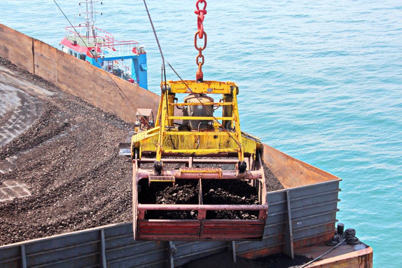 Loading coal from cargo barges onto a bulk carrier using ship cranes and grabs at the port of Samarinda, Indonesia. View of a close-up of the work of bulldozers and loaders. Loading coal from cargo barges onto a bulk carrier using ship cranes and grabs at the port of Samarinda, Indonesia. View of a close-up of the work of bulldozers and loaders.