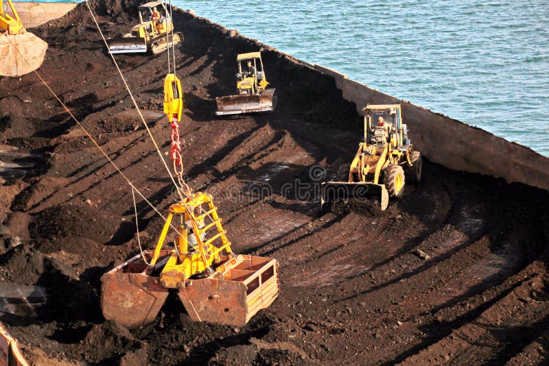 Loading coal from cargo barges onto a bulk carrier using ship cranes and grabs at the port of Samarinda, Indonesia. View of a close-up of the work of bulldozers and loaders. Loading coal from cargo barges onto a bulk carrier using ship cranes and grabs at the port of Samarinda, Indonesia. View of a close-up of the work of bulldozers and loaders.