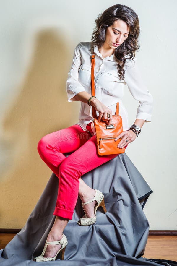 Young beautiful woman wearing white long sleeve shirt, red pants, light color sandals, shoulder carrying brown leather purse bag,  looking down, hand putting coin inside bag. Studio Shoot. Young beautiful woman wearing white long sleeve shirt, red pants, light color sandals, shoulder carrying brown leather purse bag,  looking down, hand putting coin inside bag. Studio Shoot