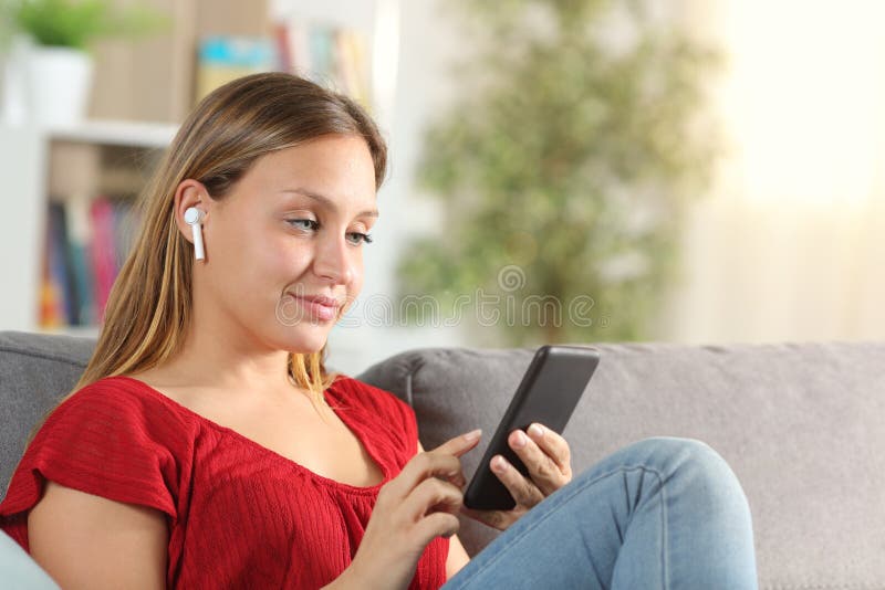 Lady listening to music with mobile phone and wireless earbuds sitting on a couch in the living room at home. Lady listening to music with mobile phone and wireless earbuds sitting on a couch in the living room at home