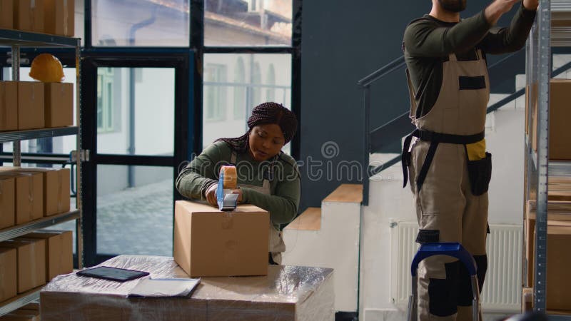 Dolly out shot of head of operations in warehouse supervising trainee sealing cardboard box parcels. Retail depository personnel properly securing goods to avoid damages during shipping. Dolly out shot of head of operations in warehouse supervising trainee sealing cardboard box parcels. Retail depository personnel properly securing goods to avoid damages during shipping