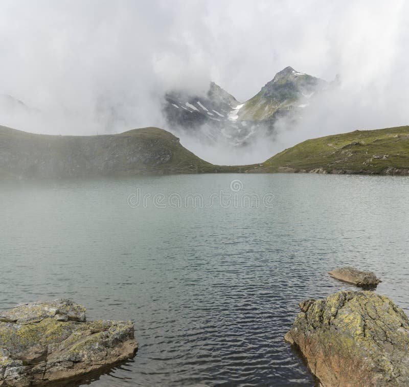 Lago idílico de la montaña con la niebla que levanta para revelar las altas montañas alpinas imagen de archivo Lago idílico de la montaña con la niebla que levanta para revelar las altas montañas alpinas imagen de archivo