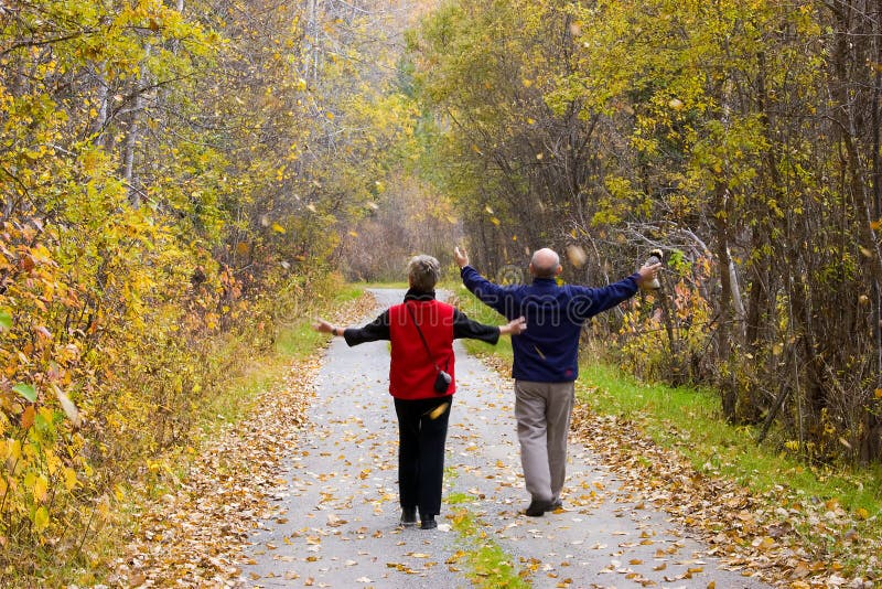 Two active seniors (mature adults) walk through falling leaves on country path with arms outstretched. Various colors of autumn leaves fall around them. Two active seniors (mature adults) walk through falling leaves on country path with arms outstretched. Various colors of autumn leaves fall around them.