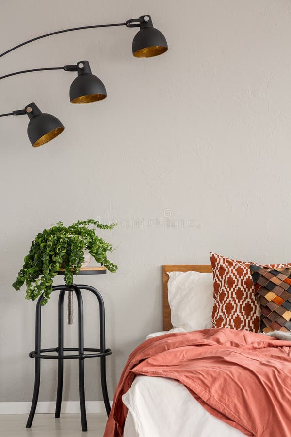 Lamp and plant on table next to red bed with pillows in grey bedroom interior