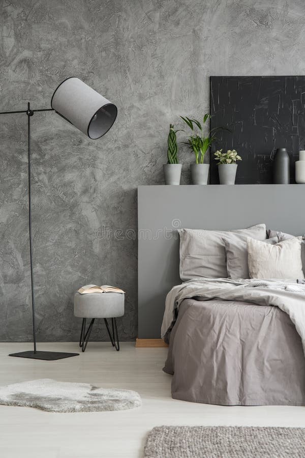 Lamp and stool next to bed with pillows and bedhead in grey bedroom interior with poster. Real photo