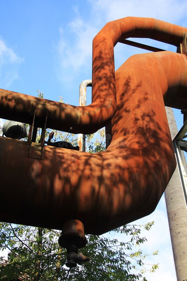 Landschaftspark Duisburgo, Alemania: Opinión de ángulo bajo sobre tubería corroída curvada aislada contra el cielo azul y árboles fotografía de archivo libre de regalías Landschaftspark Duisburgo, Alemania: Opinión de ángulo bajo sobre tubería corroída curvada aislada contra el cielo azul y árboles fotografía de archivo libre de regalías