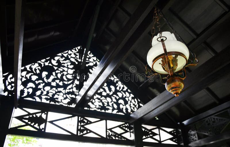 An image of a traditional ethnic design lantern or lamp hanging from the ceiling of a Malay house pavilion. Made with wrought iron metal and frosted glass. Antique design. Taken with background of carved wooden pavilion roof. Horizontal color format photograph, nobody in picture. Exotic travels photography with local art and cultural interest. An image of a traditional ethnic design lantern or lamp hanging from the ceiling of a Malay house pavilion. Made with wrought iron metal and frosted glass. Antique design. Taken with background of carved wooden pavilion roof. Horizontal color format photograph, nobody in picture. Exotic travels photography with local art and cultural interest.