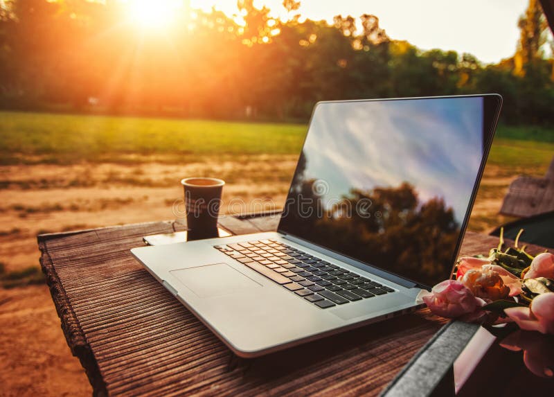 Laptop computer on rough wooden table with coffee cup and bouquet of peonies flowers in outdoor park