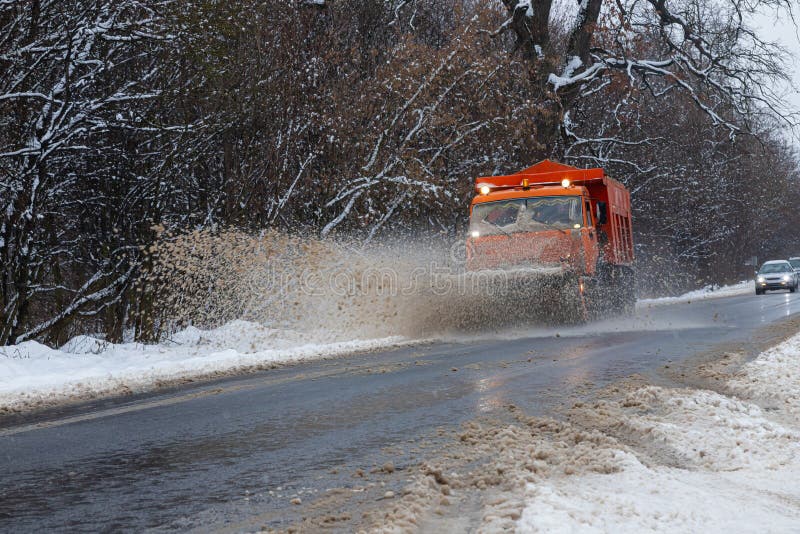 A large car with a plow clears the road from snow. Orange cargo special equipment is struggling with the elements in