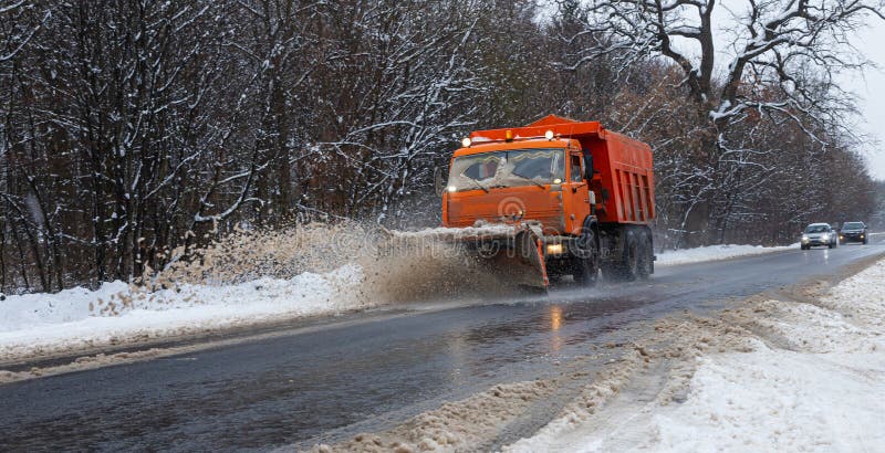 A large car with a plow clears the road from snow. Orange cargo special equipment is struggling with the elements in