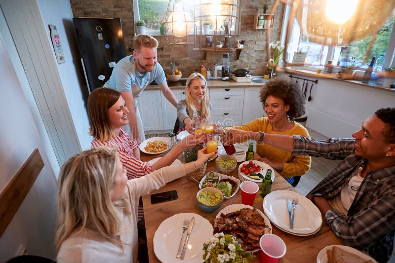 Large group of multi ethic young adults having lunch together in a domestic kitchen, toasting with drinks by the table royalty free stock photos