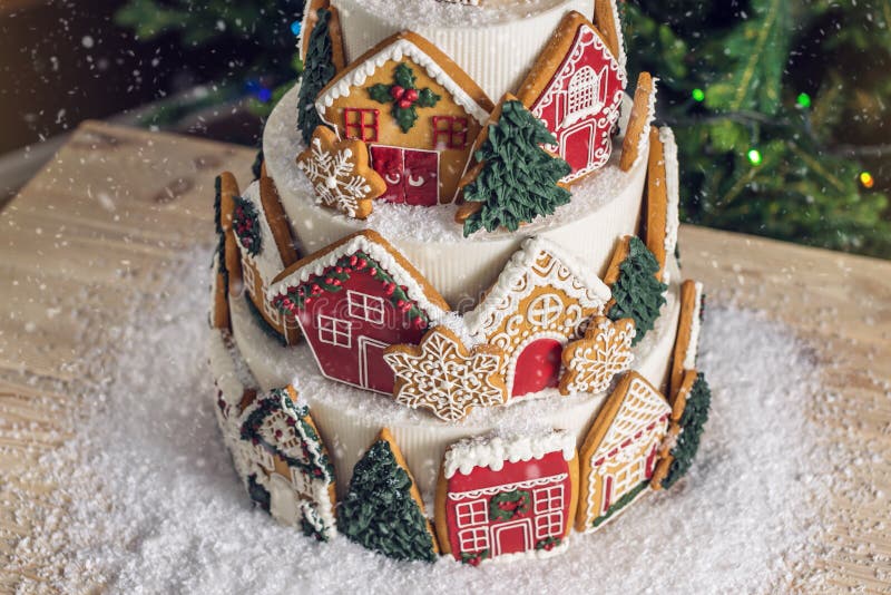 Large tiered Christmas cake decorated with gingerbread cookies and a house on top. Tree and garlands in the background. Home, ginger.