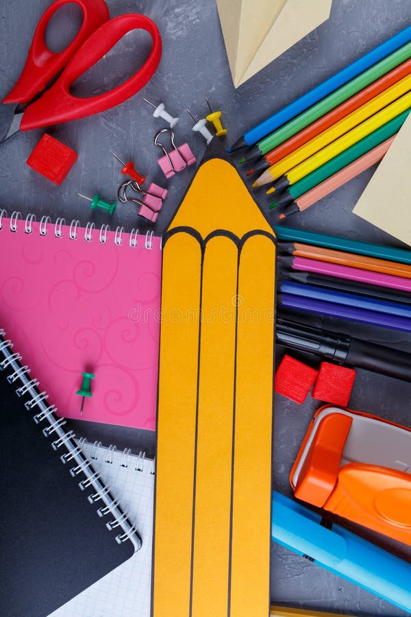 A large yellow paper pencil, next to a variety of multi-colored pencils, several notebooks, clamps and crayons and other office supplies. View from above. A large yellow paper pencil, next to a variety of multi-colored pencils, several notebooks, clamps and crayons and other office supplies. View from above.