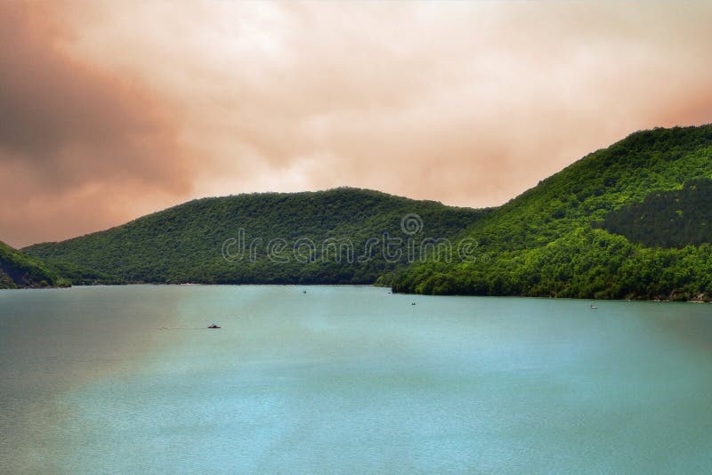 Las montañas con el bosque verde en un lago apuntalan en el cielo tempestuoso amarillo con el fondo de las nubes fotografía de archivo libre de regalías Las montañas con el bosque verde en un lago apuntalan en el cielo tempestuoso amarillo con el fondo de las nubes fotografía de archivo libre de regalías