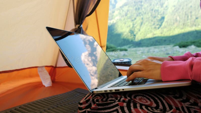 Las montañas se reflejan en la pantalla del ordenador portátil La mujer del Freelancer utiliza el ordenador en una tienda turísti