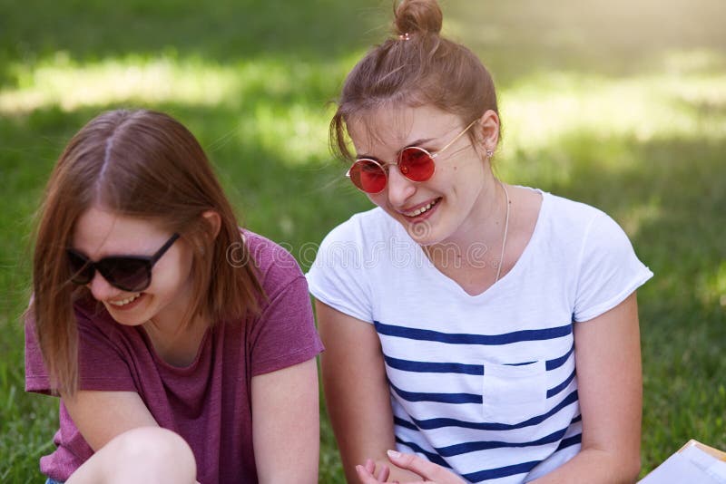 Las mujeres felices que hablan y que ríen en el parque, sentándose en hierba, llevan la ropa casual y las gafas de sol, oyen brom fotos de archivo libres de regalías Las mujeres felices que hablan y que ríen en el parque, sentándose en hierba, llevan la ropa casual y las gafas de sol, oyen brom fotos de archivo libres de regalías