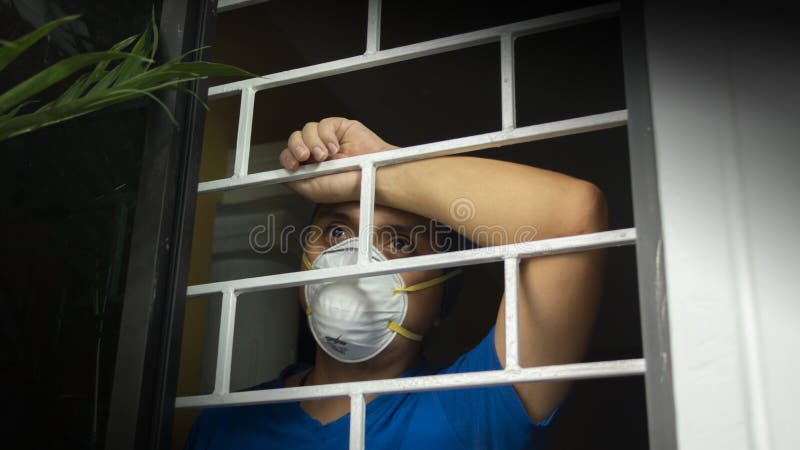 Latin man with white face mask inside his house looking out through the window, leaning against security bars