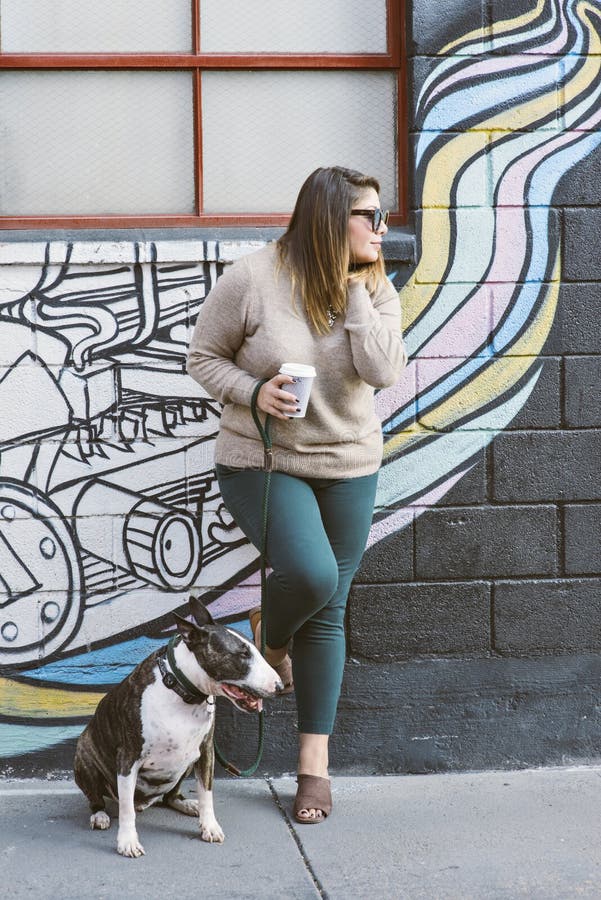 Latina Millennial stojaki Przeciw ścianie z Jej zwierzę domowe psa Bull Terrier obsiadaniem Jej stroną fotografia stock Latina Millennial stojaki Przeciw ścianie z Jej zwierzę domowe psa Bull Terrier obsiadaniem Jej stroną fotografia stock