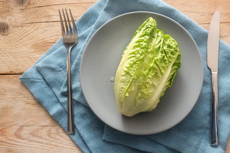 Fresh green lettuce or baby cos on a gray plate with cutlery on a blue napkin and rustic wooden planks, concept for a healthy vegetarian diet to lose weight, copy space, high angle view from above. Fresh green lettuce or baby cos on a gray plate with cutlery on a blue napkin and rustic wooden planks, concept for a healthy vegetarian diet to lose weight, copy space, high angle view from above