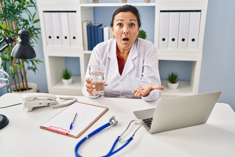 Hispanic mature doctor woman holding pill and glass of water afraid and shocked with surprise and amazed expression, fear and excited face. Hispanic mature doctor woman holding pill and glass of water afraid and shocked with surprise and amazed expression, fear and excited face