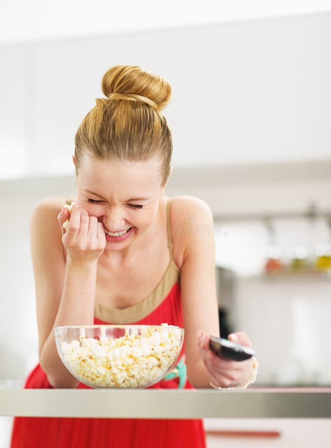Laughing young woman eating popcorn and watching tv in kitchen stock photo Laughing young woman eating popcorn and watching tv in kitchen stock photo