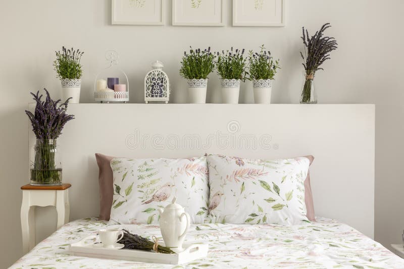 Lavender bedroom interior with a bed, pillows, wooden tray with pot, flowers and decorations. Real photo