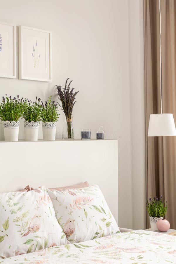 Lavender flowers on bedhead above bed with pillows in bright bedroom interior with posters and lamp. Real photo concept