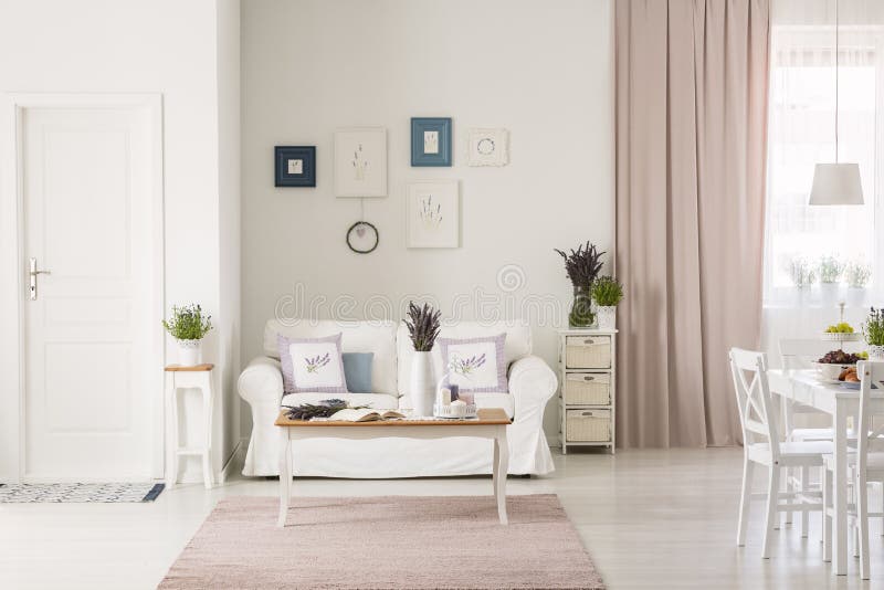 Lavender flowers on table in front of white couch in flat interior with pink drapes and posters. Real photo concept