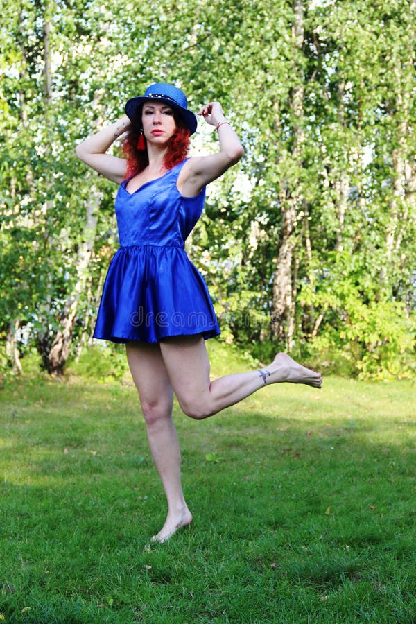 Woman on the lawn with red curly hair in a blue hat and short blue mini dress. Holding a hat. stands on one leg. royalty free stock image Woman on the lawn with red curly hair in a blue hat and short blue mini dress. Holding a hat. stands on one leg. royalty free stock image