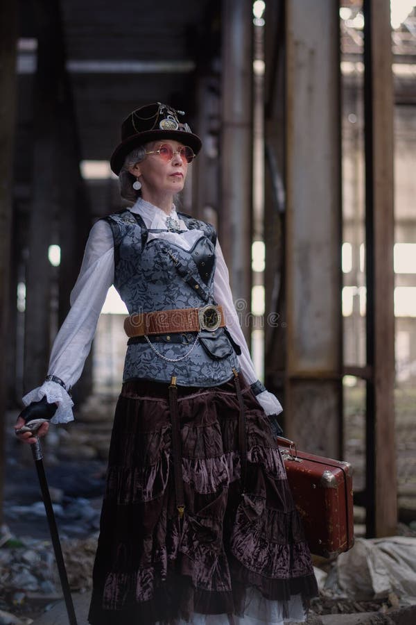 ?ldre dam i en steampunkdr?kt p? en ?vergiven fabrik med armar i hand arkivfoto ?ldre dam i en steampunkdr?kt p? en ?vergiven fabrik med armar i hand arkivfoto